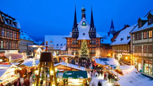 Ein verschneiter Weihnachtsmarkt in Deutschland mit beleuchteten Buden, einem Weihnachtsbaum und einem historischen Rathaus im Hintergrund.